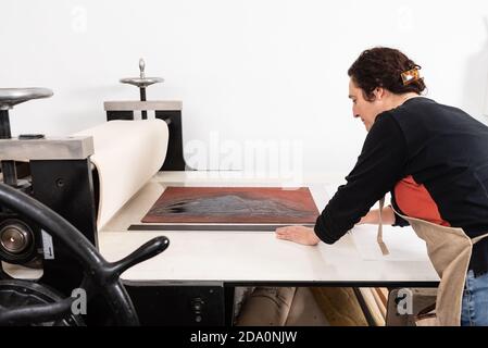 Focalizzato medio invecchiato femmina master mettendo foglio di linoleum con ornamento inchiostrato sulla pressa da stampa mentre si lavora in tecnica di linocut in studio Foto Stock