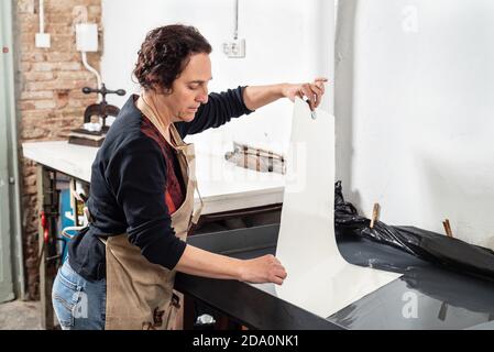 Vista laterale di messa specializzata artista femminile di mezza età messa a fuoco foglio di carta sulla pressa di stampa durante la creazione di grafica in stampa tecnica in profes Foto Stock