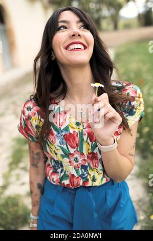 Positiva giovane femmina in abbigliamento casual brillante guardando in su con sorrisi luminoso mentre tendi piccolo mentre delicato daisy nel cortile su sfondo sfocato Foto Stock