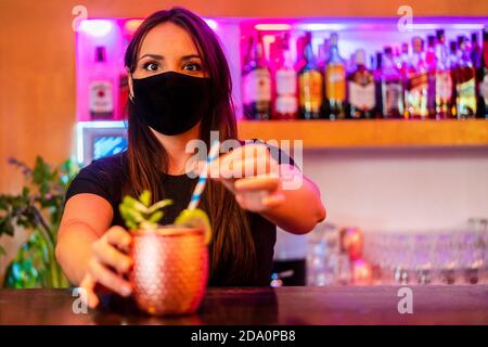 Giovane barattiera femminile in maschera protettiva che serve cocktail con paglia mentre si lavora in bar Foto Stock