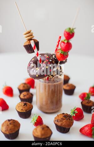 Bicchiere di frullato di cioccolato guarnito con fragole fresche e assortito pasticceria dolce su tavola bianca Foto Stock