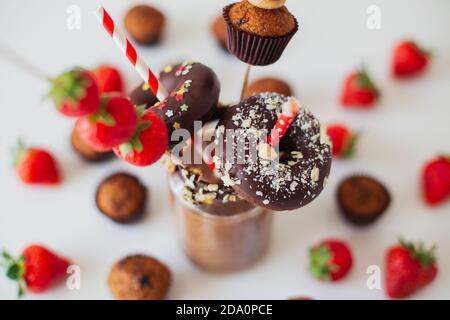 Bicchiere di frullato di cioccolato guarnito con fragole fresche e assortito pasticceria dolce su tavola bianca Foto Stock