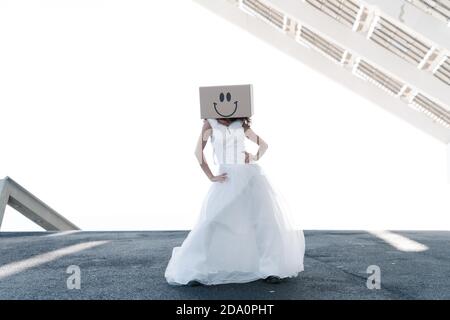 Corpo pieno di donna anonima in abito da sposa con sorridente scatola sulla testa in abito da sposa che tiene le mani sulla vita sotto il tetto dell'edificio locale Foto Stock