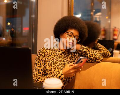 Concentrato afroamericano femmina di navigazione telefono cellulare durante il lavoro online in caffetteria vicino allo specchio Foto Stock