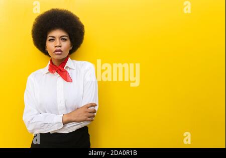 Donna afro-americana sicura in un elegante abito formale con cui stare in piedi braccia incrociate su sfondo giallo Foto Stock