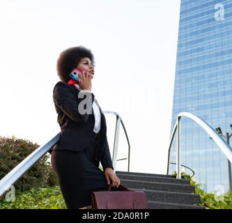 Vista laterale ad angolo basso di una seria imprenditrice afroamericana in un abbigliamento formale con valigia portadocumenti con telefonata in strada Foto Stock