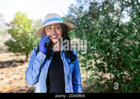 Sorridente giovane donna che parla al telefono mentre lavora in verde giardino con alberi da frutta Foto Stock