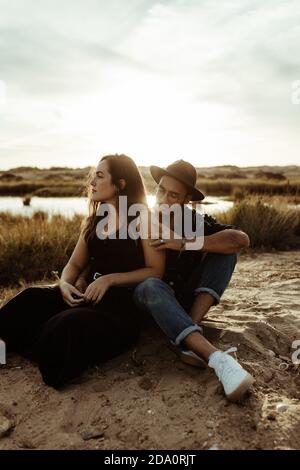 Giovane coppia amorosa in abbigliamento casual seduta insieme guardando via sulla costa di bacino idrico circondato da erba secca in prato alla luce del sole Foto Stock