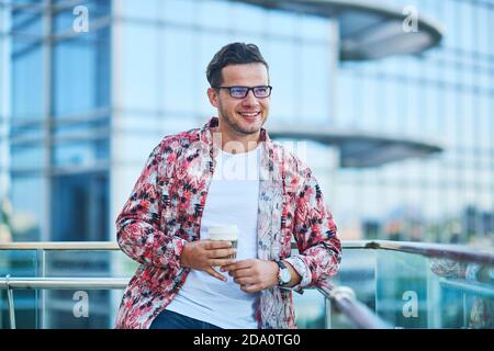 Giovane uomo sorridente in abito alla moda con ornamento in piedi con bicchiere di plastica di bevanda calda guardando lontano vicino all'edificio Foto Stock