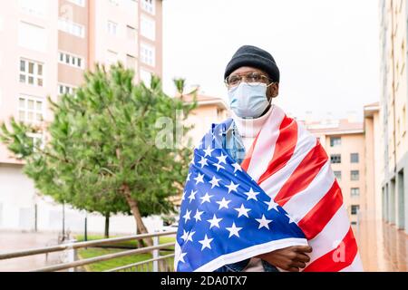 Uomo patriottico afroamericano in maschera medica avvolta negli Stati Uniti bandiera nazionale in piedi sulla strada durante la pandemia di coronavirus e guardare via Foto Stock