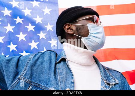 Uomo patriottico afroamericano in maschera medica avvolta negli Stati Uniti bandiera nazionale in piedi sulla strada durante la pandemia di coronavirus e guardare via Foto Stock