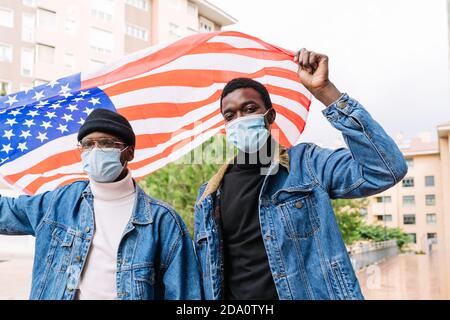Basso angolo di allegri amici africani americani maschi in maschere In piedi con la bandiera nazionale degli Stati Uniti sulla strada ed esprimendo gioia Foto Stock