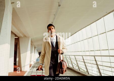 Uomo serio in abiti eleganti con valigia che cammina lungo il corridoio all'aeroporto di giorno Foto Stock