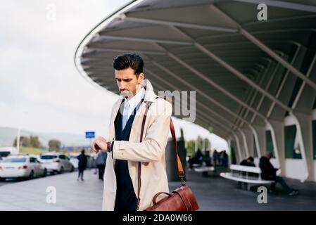 uomo con capelli scuri in tuta e cappotto guardando il tuo orologio mentre si trova vicino al terminal dell'aeroporto di giorno Foto Stock