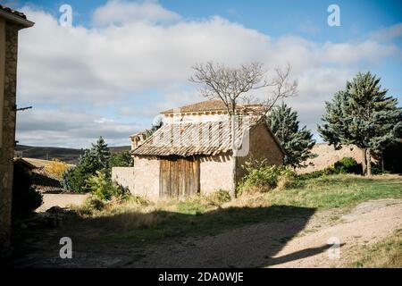 Edificio in pietra intemperie con tetto in tegole di shabby e senza foglie essiccate albero che cresce nelle vicinanze in villaggio situato su terreno collinare con alberi verdi in soleggiato a Foto Stock