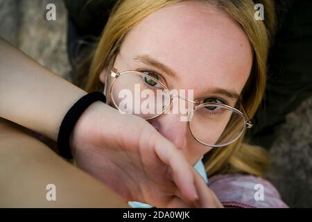 Crop tranquilla giovane femmina con capelli biondi in occhiali eleganti inclinati a portata di mano e guardando la fotocamera mentre si riposa in natura Foto Stock