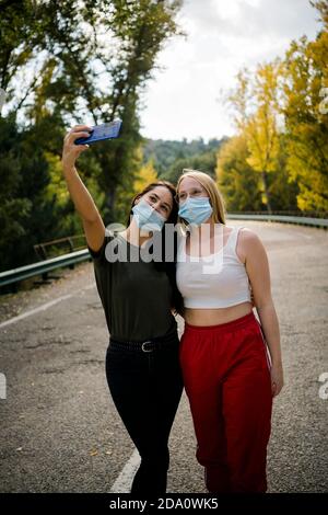 Allegra giovane amica in abiti casual e maschere protettive prendere selfie su smartphone mentre si è in piedi su strada asfaltata che passa attraverso la foresta Foto Stock