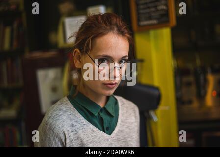 Donna intelligente seria che indossa abiti casual e occhiali in piedi accogliente biblioteca sullo sfondo di librerie e guardando la macchina fotografica Foto Stock