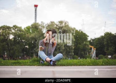 Giovane studentessa con capelli rossi in elegante abito casual e. cappello con smartphone e ascolto di musica e relax parco estivo Foto Stock