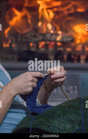 Mani di donna che si maglia e stufa a cassetta che brucia sullo sfondo. Foto Stock