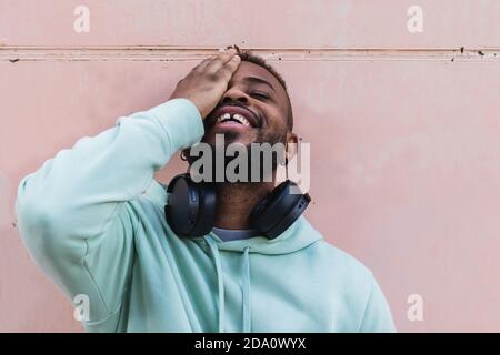 Allegro ragazzo alato afroamericano adulto in abbigliamento casual con cuffie sul collo che coprono l'occhio contro le pareti grigie Foto Stock