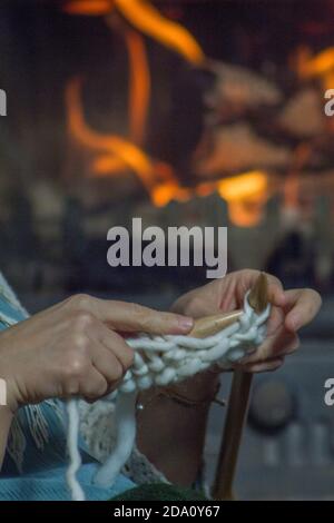 Primo piano di mani femminili e aghi da maglieria con camino sullo sfondo. Foto Stock