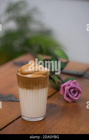 Bicchiere di delizioso caffè fresco Dalgona posto su un tavolo di legno con fiori di rosa al caffè Foto Stock
