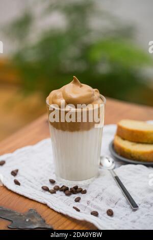 Bicchiere di gustosa bevanda Dalgona con caffè istantaneo montato e. latte messo sul tavolo nel caffè con torta per la colazione Foto Stock