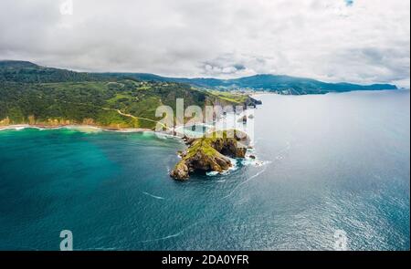 Vista drone della pavimentazione in pietra che conduce lungo il ponte di pietra e cresta di collina rocciosa a casa solitaria sull'isola Gaztelugatxe circondato da mare tranquillo wa Foto Stock