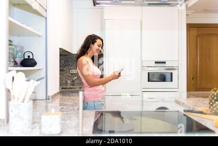 Vista laterale di una giovane donna felice in abiti casual in piedi vicino al bancone della cucina e chiacchierando con il telefono cellulare durante la spesa tempo a casa Foto Stock
