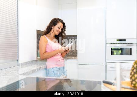 Vista laterale di una giovane donna felice in abiti casual in piedi vicino al bancone della cucina e chiacchierando con il telefono cellulare durante la spesa tempo a casa Foto Stock