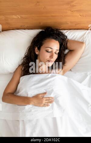 Da sopra femmina calmo in pajama sdraiato a letto sopra morbido cuscino sotto coperta bianca e sonno dolce Foto Stock