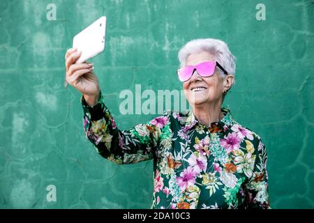 Felice femmina con capelli grigi invecchiati in outfit e occhiali da sole eleganti prendere selfie sul telefono cellulare mentre si sta in piedi contro parete verde Foto Stock