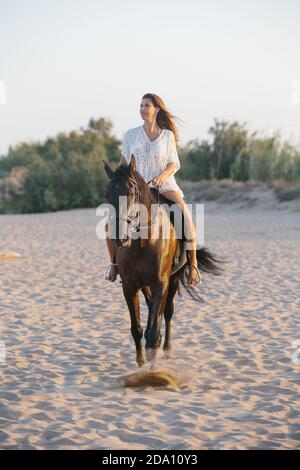 Bella donna in vestito bianco cavalcando un cavallo sulla spiaggia al tramonto Foto Stock