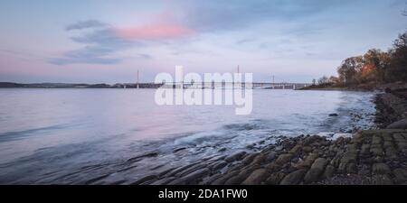 Panorama della costa della baia del mare con pietre In primo piano al tramonto vicino al New Queensferry Crossing ponte in Scozia Foto Stock