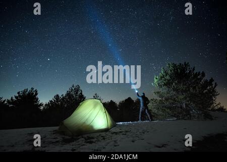 Uomo in piedi vicino alla tenda illuminata e puntando la torcia blu al cielo stellato in inverno Nebrodi Park, Sicilia Foto Stock