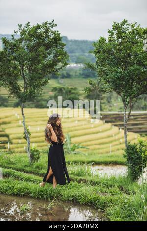 Donna che cammina contro le fantastiche terrazze di riso di Jatiluwih a Bali. Bellezza e armonia nella natura. Indossabile in un lungo abito nero. Foto Stock