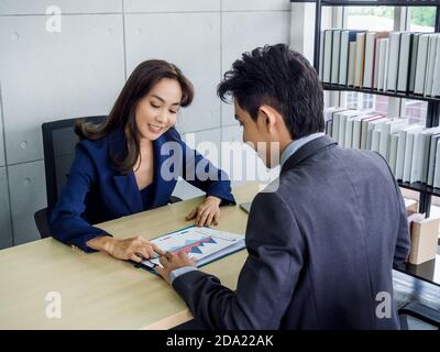 Donna asiatica d'affari e uomo d'affari che lavorano insieme in ufficio. La boss femminile spiega il grafico a un giovane dipendente di sesso maschile che indossa il vestito sulla scrivania. Foto Stock