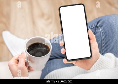 Immagine mockup di una donna che tiene e utilizza il telefono cellulare nero con schermo da tavolo vuoto mentre si beve caffè in caffetteria. Mano di donna che tiene in mano mobil bianco Foto Stock