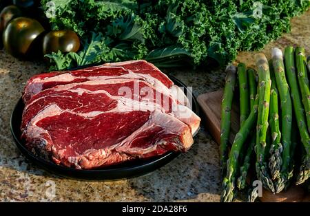 Carne cruda, bistecca di manzo su piatto nero con kale, asparagi, broccolini e pomodori sullo sfondo. Foto Stock