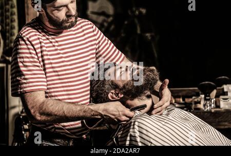 Il barbiere rasa la barba. Un uomo con la bearded seduto in una poltrona in un barbiere mentre un parrucchiere rasa la barba con un rasoio pericoloso Foto Stock