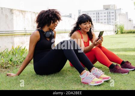 Gruppo vario di due amici di idoneità femminili che parlano e usando un telefono mobile nel parco. Giovani donne che riposano e sorridono all'aperto Foto Stock