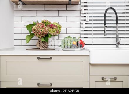 Mobili da cucina bianchi con maniglie in metallo sulle porte vicino al lavabo con un bouquet di fiori e un piatto di verdure. Foto Stock