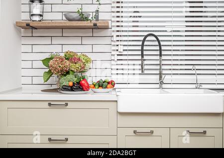 Mobili da cucina bianchi con maniglie in metallo sulle porte vicino al lavabo con un bouquet di fiori e un piatto di verdure. Foto Stock