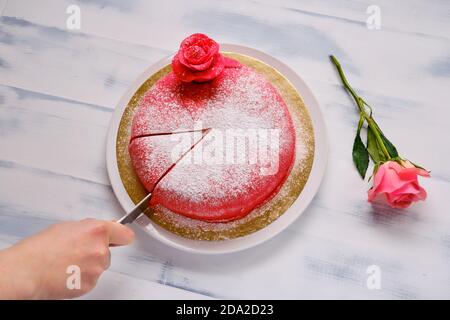 Una mano con un coltello taglia un pezzo da una torta rossa. Tavolo di legno bianco con rosa rossa Foto Stock