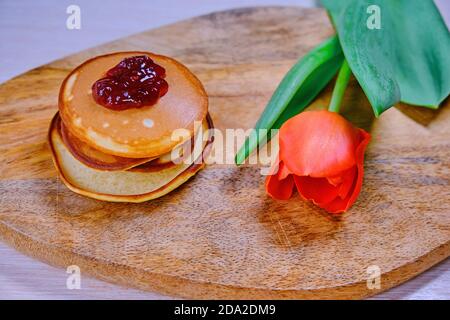 Red Tulip accanto a una pila di pancake. Torte calde fatte in casa su un tavolo di legno. Marmellata di frutti di bosco su frittelle e fiori viola. Foto Stock