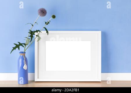 Elegante cornice orizzontale bianca con mockup opaco, modello con lancette a globo in vaso davanti alla parete blu chiaro. Area immagine vuota isolata Foto Stock