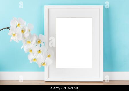 Elegante modello mockup con cornice verticale bianca con opaco, e orchidea in fiore ramo di fronte a parete blu pastello. ima vuoto Foto Stock