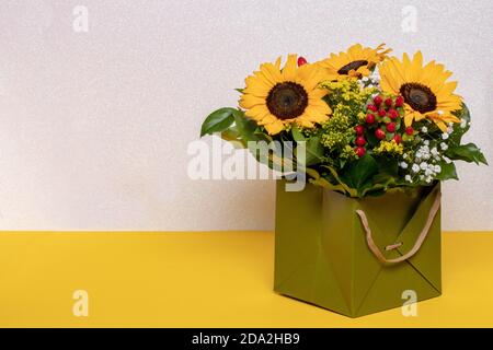 Closeup di un bel bouquet con girasoli gialli in una scatola regalo decorativa verde su un tavolo giallo su uno sfondo rosa chiaro scintillante. Macro. Foto Stock