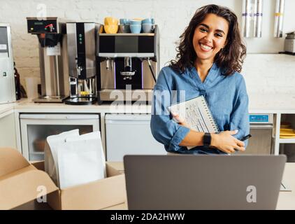 Ritratto di una felice proprietaria di una caffetteria. Donna d'affari che tiene un notebook nel caffè e guarda la macchina fotografica. Foto Stock